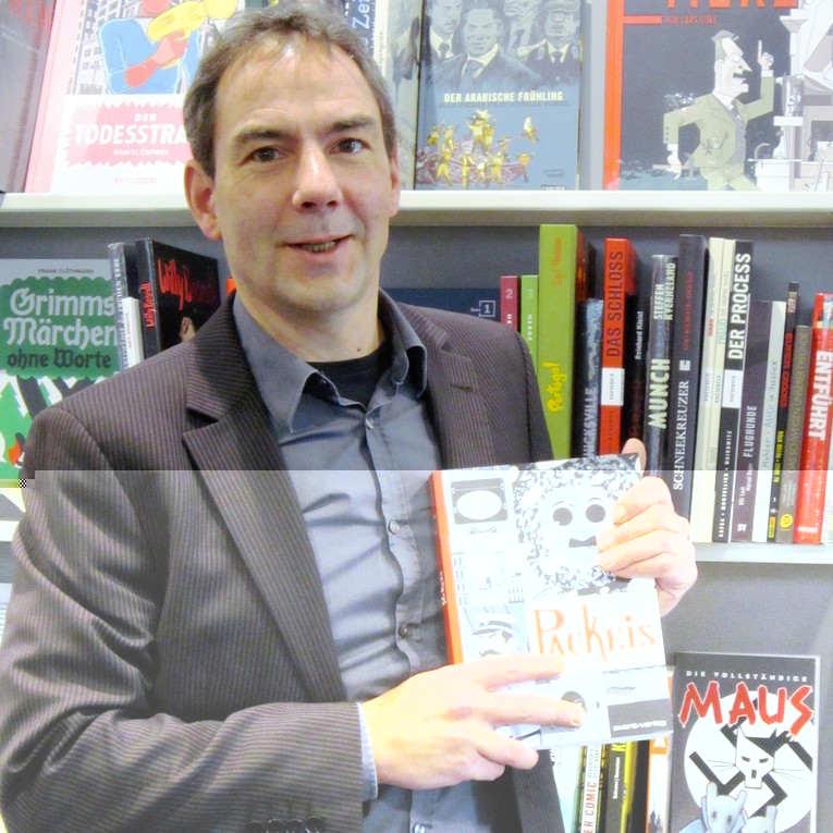 Volker Petri steht vor dem Comic-Regal, in seinen Händen die Graphic Novel "Packeis"