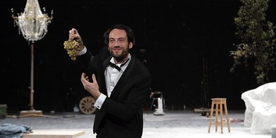 Szenenfoto 'Crocodile Trompeur': Ein lächelnder Mann im Frack hält mit der rechten Hand eine Rebe Weintrauben und verweist mit der linken darauf. Im Hintergrund ein Kronleuchter, ein Hocker und andere Gegenstände.
