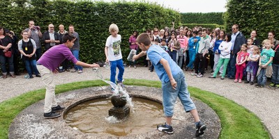 Drei Jungen vollführen Wasserspiele im Großen Garten in Herrenhausen. Zahlreiche Kinder und Erwachsene im Hintergrund beobachten sie dabei.