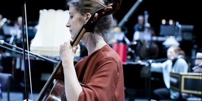 Szenenfoto 'Songs of War': Im Bild eine Cello spielende Frau. Im Hintergrund verschwommen weitere Orchestermitglieder.